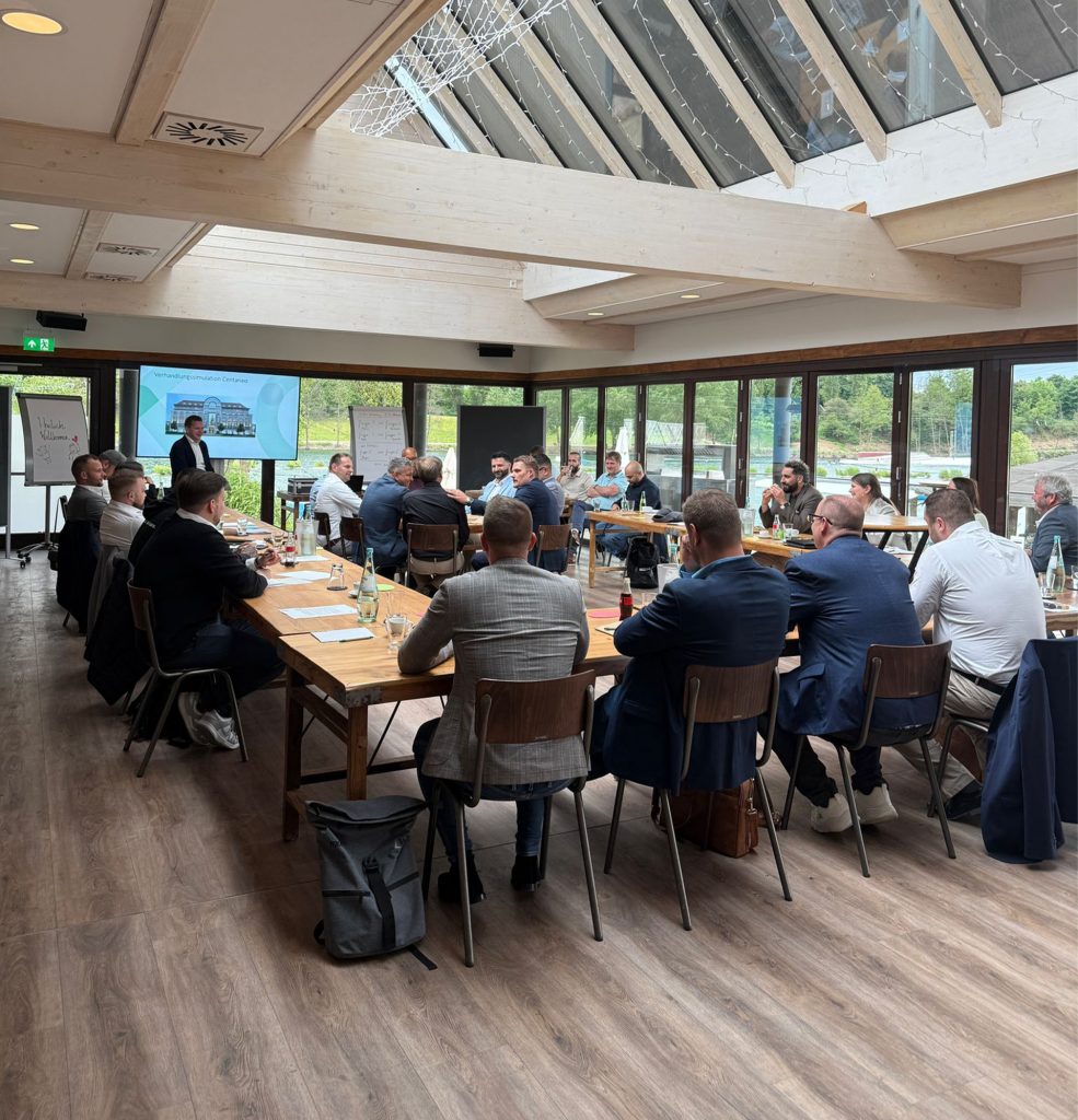 Das Bild zeigt einen Konferenzraum, wo unsere Vertriebler an einem U-förmigen Tisch sitzen und gerade eine Vertriebssimulation durchspielen. Dazu wurde in der Mitte des Raumes ein Tisch gestellt und ein Vertriebsgespräch simuliert. Das Bild entstand während der Vertriebsschulung.